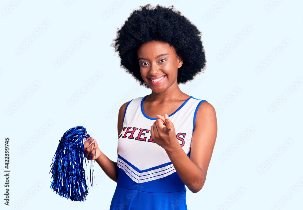 Sticker Young african american woman wearing cheerleader uniform holding pompom beckoning come here gesture with hand inviting welcoming happy and smiling