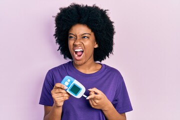 Young african american woman holding glucometer device angry and mad screaming frustrated and furious, shouting with anger. rage and aggressive concept.