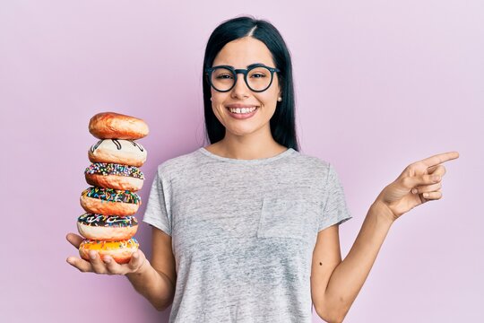 Beautiful young woman holding pile of tasty colorful doughnuts smiling happy pointing with hand and finger to the side