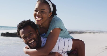 African american man smiling and carrying african american woman piggyback on the beach - Powered by Adobe