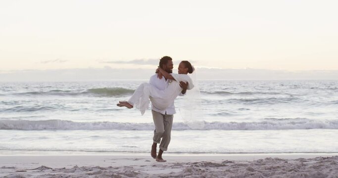 African american couple in love getting married, man carrying woman on the beach at sunset