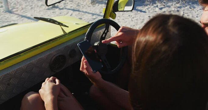 Happy caucasian couple sitting in beach buggy by the sea using smartphone