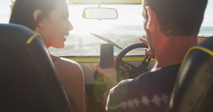 Happy caucasian couple sitting in beach buggy by the sea using smartphone