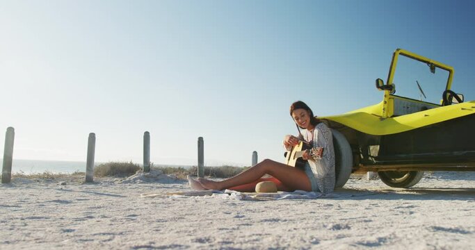 Happy caucasian woman sitting beside beach buggy by the sea playing guitar