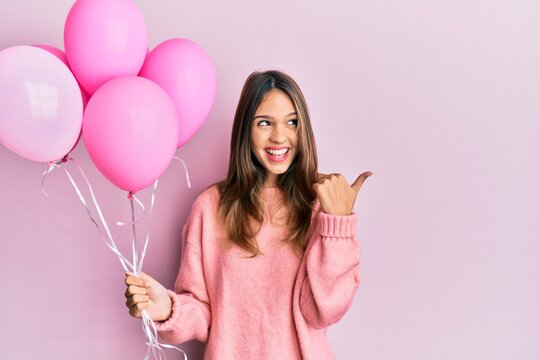 Young brunette woman holding pink balloons pointing thumb up to the side smiling happy with open mouth