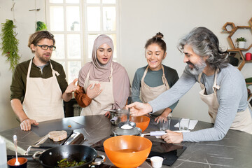 Happy mature bearded man holding kitchen tool over metallic pan with boiling water while cooking by table among learners of masterclass