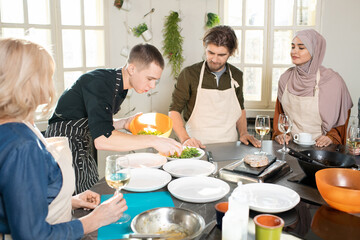 Young male cooking coach in apron bending over large kitchen table and taking fresh ruccola for salad among learners during masterclass