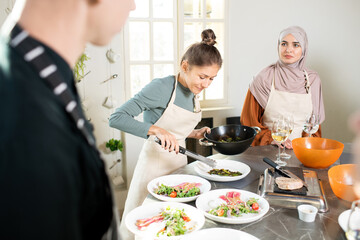 Young pretty blond female with frying pan bending over large kitchen table while putting vegetable stew on plate before serving it