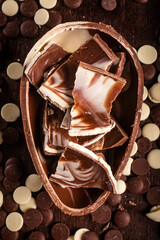 Top view of dark and white chocolate easter egg with cracked eggs inside and lots of chocolate chips all around on a wooden table.