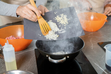 Hands of mature man with wooden spatula putting chopped onions into hot frying pan with oil while other trainee standing next to him
