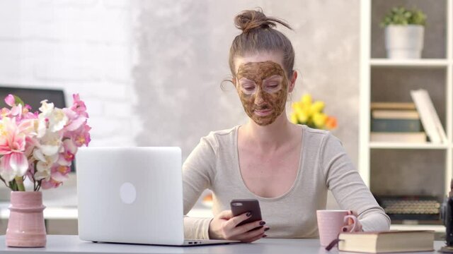 Woman cosmetic facial skin care. Woman in skin rubber peeling face beauty mask sitting at desk using phone and laptop.