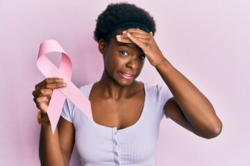 Young african american girl holding pink cancer ribbon stressed and frustrated with hand on head,...