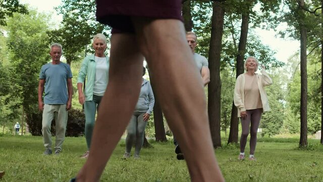 Wide shot of group of five aged sporty people doing forward lunges in park while unrecognizable trainer watching them