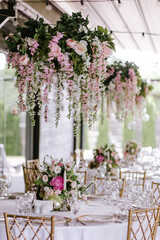 Fototapeta na wymiar Gold chairs and tables decorated with natural pink and green flowers, candles and gold frames. Hanging flower beds and light bulbs Wedding decoration and decor, floristic concept