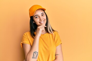 Young hispanic woman wearing delivery uniform and cap looking confident at the camera with smile with crossed arms and hand raised on chin. thinking positive.