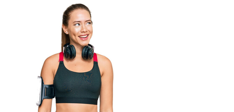 Beautiful blonde woman wearing gym clothes and using headphones looking away to side with smile on face, natural expression. laughing confident.