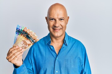 Middle age bald man holding canadian dollars looking positive and happy standing and smiling with a confident smile showing teeth