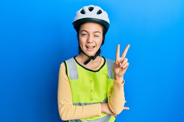 Beautiful brunette little girl wearing bike helmet and reflective vest smiling with happy face winking at the camera doing victory sign. number two.