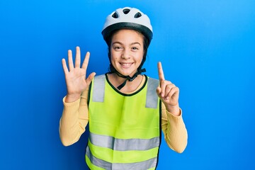 Beautiful brunette little girl wearing bike helmet and reflective vest showing and pointing up with fingers number six while smiling confident and happy.