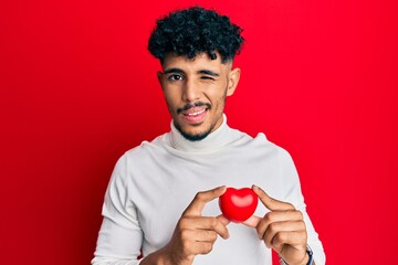 Young arab handsome man holding heart winking looking at the camera with sexy expression, cheerful...