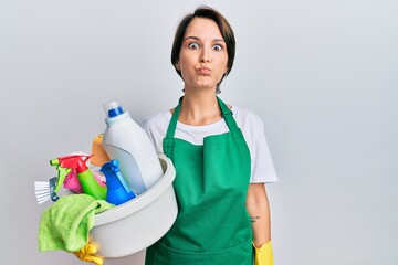 Young brunette woman with short hair wearing apron holding cleaning products puffing cheeks with funny face. mouth inflated with air, crazy expression.