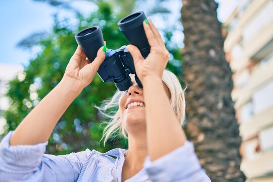 Young blonde woman smiling happy looking for new opportunity using binoculars at the park,