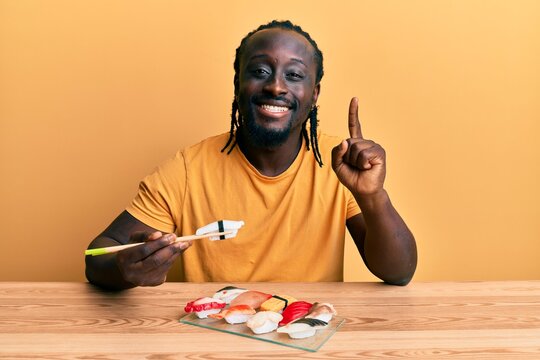 Handsome young black man eating sushi sitting on the table smiling with an idea or question pointing finger with happy face, number one