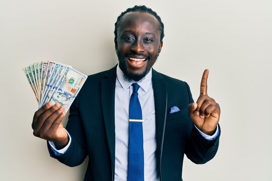 Handsome young black man wearing business suit and tie holding dollars smiling with an idea or question pointing finger with happy face, number one