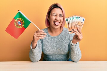 Hispanic woman with pink hair holding portugal flag and euros banknotes winking looking at the...