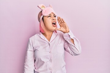 Hispanic woman with pink hair wearing sleep mask and pajama shouting and screaming loud to side with hand on mouth. communication concept.
