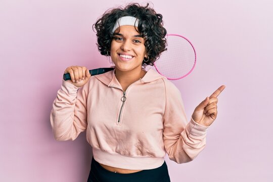 Young hispanic woman with curly hair holding badminton racket smiling happy pointing with hand and finger to the side