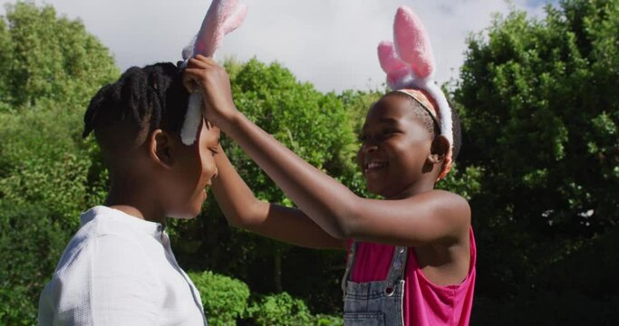 Smiling african american girl wearing easter bunny ears putting ears on her brother in garden