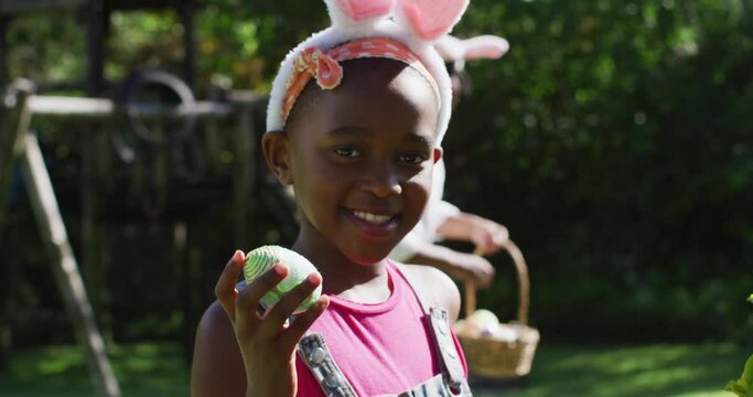African american girl wearing easter bunny ears doing easter egg hunt in garden with brother