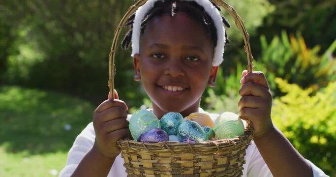 Smiling african american girl wearing easter bunny ears holding basket of easter eggs in garden