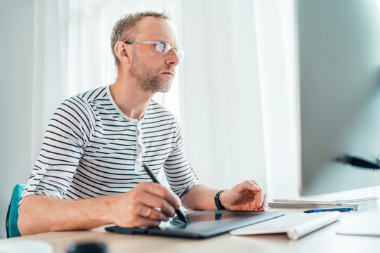 A middle-aged man in eyeglasses using a graphic tablet with pen and modern computer drawing artwork in his living room. Home office, distance or freelance work on worldwide quarantine time concept