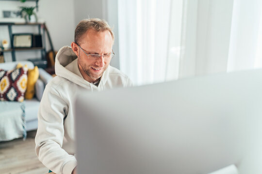 Smiling Middle-aged man in eyeglasses using a modern computer in his living room. Writing or Distance or freelance work on worldwide quarantine time concept