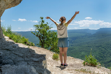 Young female stand on cliff on peak in high mountains with hands up expressing feeling of freedom and relexation. Healthy lifestyle, adventure during hiking in mountains.
