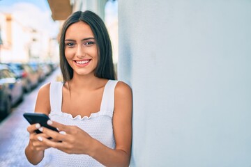 Young latin girl smiling happy using smartphone leaning on the wall.