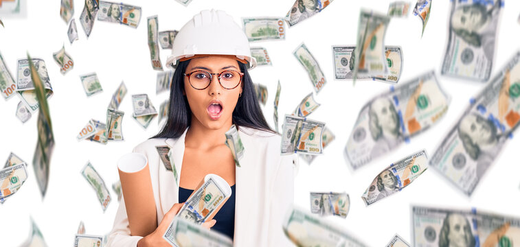 Young beautiful latin girl wearing architect hardhat holding blueprints scared and amazed with open mouth for surprise, disbelief face