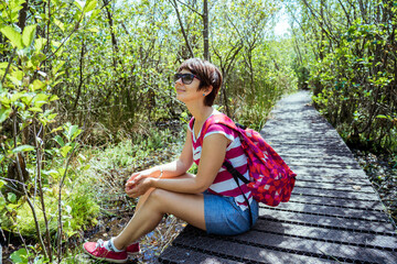 A woman with backpack sitting on the wooden pathway and relaxing after hiking in the summer forest. Enjoying, unity with nature, digital detox, personal fulfillment. Local traveling in spring, summer.