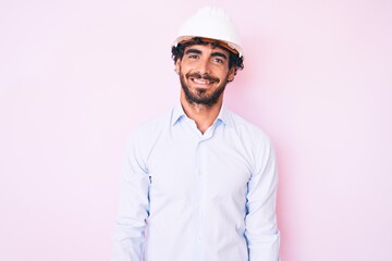 Handsome young man with curly hair and bear wearing architect hardhat with a happy and cool smile on face. lucky person.