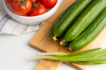 Tomatoes on a light background in a white bowl, fresh cucumbers and green onions on a cutting board.