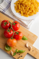 On a cutting board, a branch with tomatoes, basil leaves and garlic cloves, a bowl of pasta on a light background.