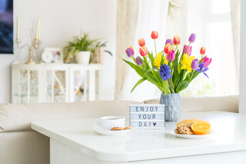 Good morning concept. Romantic breakfast - fresh spring flowers, cup of hot coffee drink, cookies, orange, lightbox with message Enjoy your day on marble table with light interior view. Copy space.