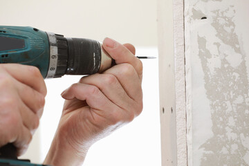 The installer worker in safe medical mask and overalls fixes a detail of the stiffener element with screws using an electric screwdriver to the wall of the metal structure