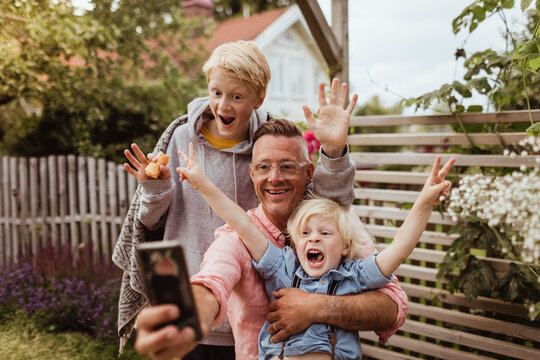 Cheerful father taking selfie with sons on smart phone in front yard at evening