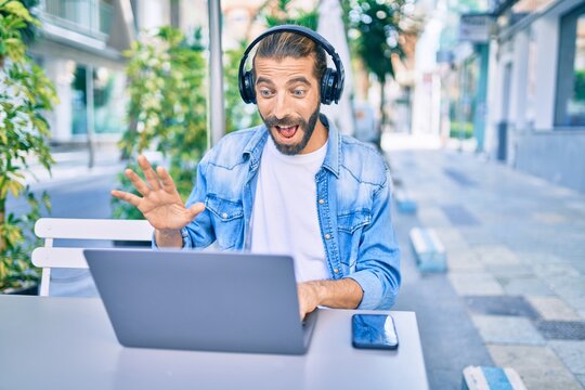 Young middle eastern man doing video call using laptop and headphones at coffee shop terrace.