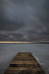 gloomy severe winter vertical gray landscape. an old wooden boardwalk pier on a large frozen lake under a thick dramatic cloudy sky. bad weather