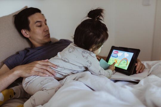 Male toddler watching cartoon on digital tablet with father in bedroom