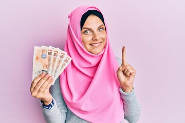 Young caucasian woman wearing islamic hijab holding 10 pounds banknotes smiling with an idea or question pointing finger with happy face, number one